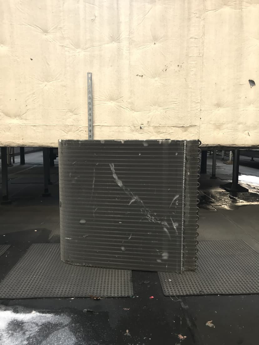 A large, coiled heat exchanger for a cooling unit replacement is placed on a rubber mat beside a beige wall on an industrial rooftop.